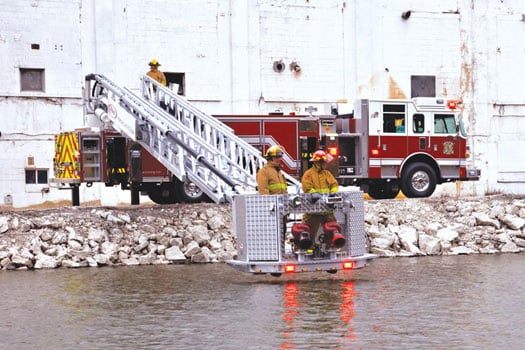 Two firefighters in the bucket of a 100’ Aluminum Aerial Platform extended out above a body of water. 