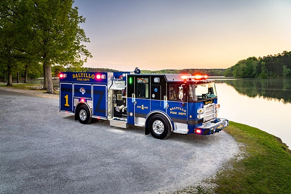 A blue Pierce Saber Pumper parked by a lake on gravel with sunset behind it.
