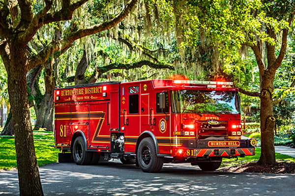 A Pierce Enforcer PUC Pumper parked under green trees on a paved road in front of green grass.