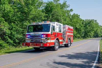 The front drivers side of a Velocity Pumper driving on a road next to a treeline on a sunny day with the lights on.