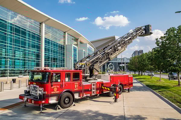 An Enforcer™ Ascendant® 100' Heavy-Duty Aerial Tower in front of a glass building with the aerial extended up on a sunny day with clouds in the sky.