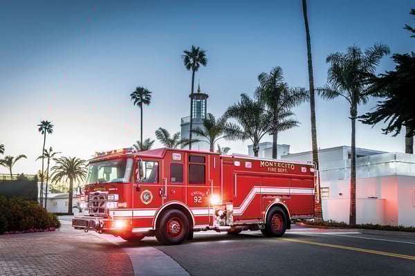 The front driver's side of an Enforcer PUC Pumper parked on a road with the lights on in front of palm trees.