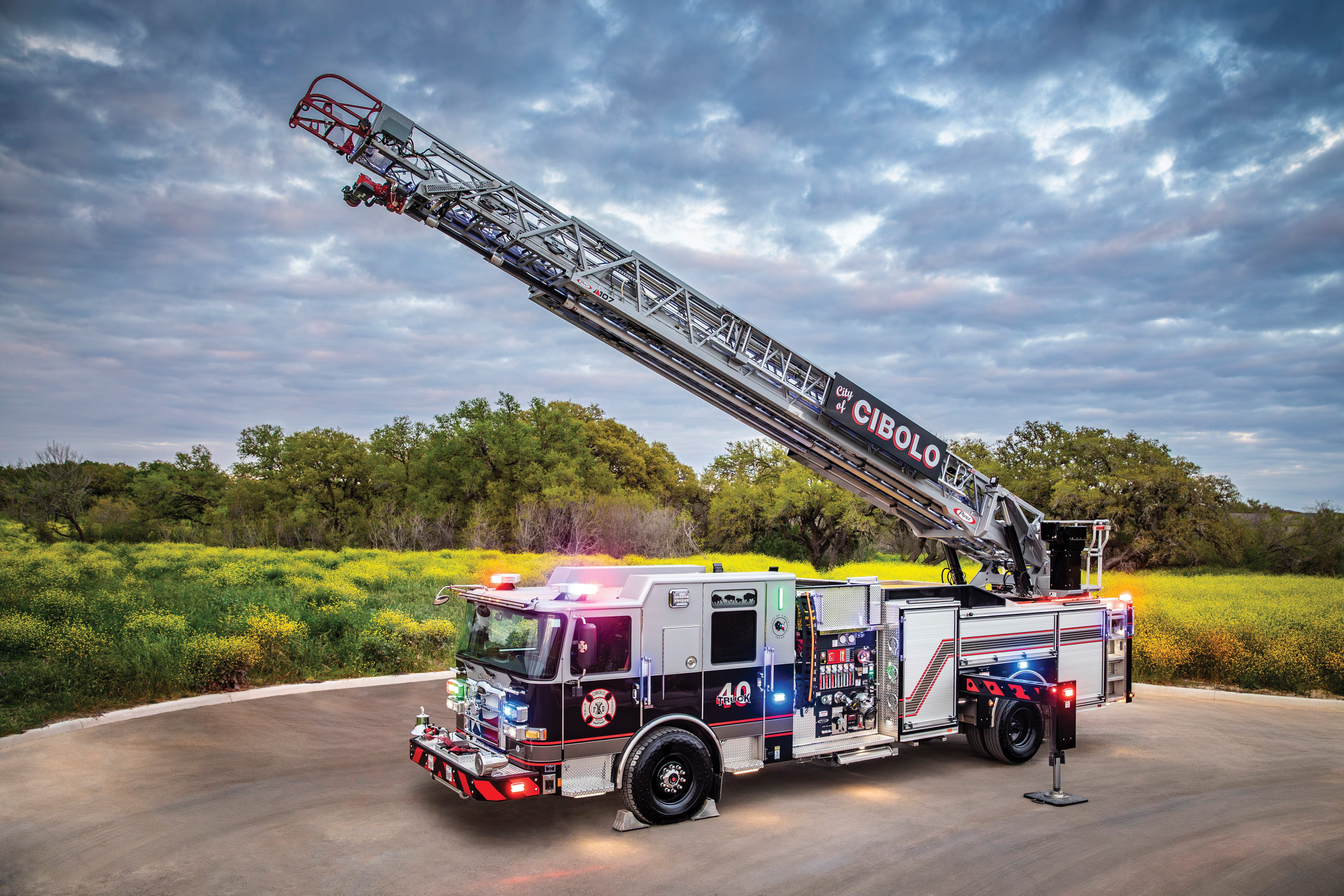Download An Enforcer Ascendant 107 Aerial Ladder with the aerial extended and lights on in front of a green grass field on a cloudy day