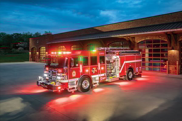 The front driver's side of a fire truck with the lights on parked in front of a fire station at night.