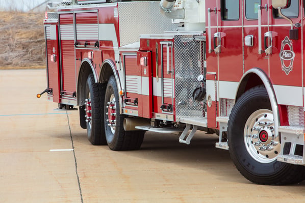 A close-up side view of a red and silver fire truck showing its large tires, the side of the truck's body, and the diamond plate compartments.