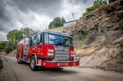 A fire truck driving on a narrow pavement road beside a steep rocky cliff on a cloudy day.