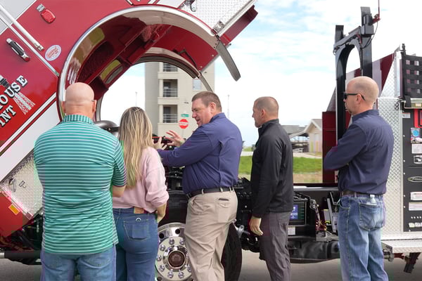 A fire truck sales representative providing customer support to firefighters next to a fire truck with the cab tilted.