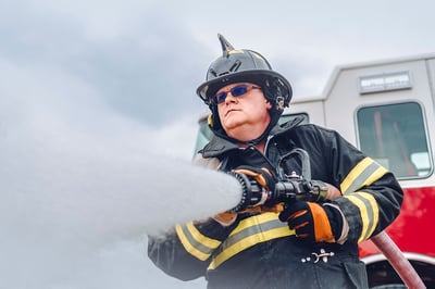 A firefighter in turnout gear spraying foam from a fire hose with a fire truck in the background.