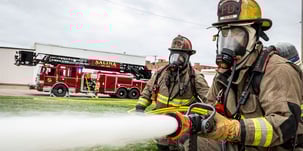 Two firefighters in turnout gear spraying water from a fire hose from a fire truck with a firefighter operating the pump.