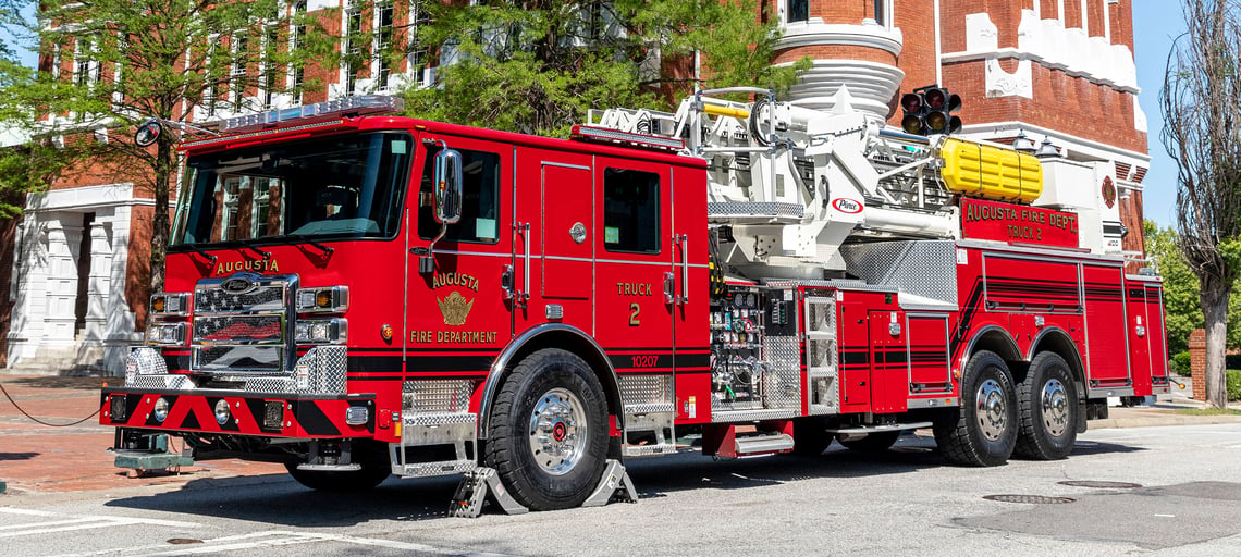 The front driver's side of an Ascendant 100' Heavy-Duty Aerial Tower parked beside a red brick building on a sunny day.
