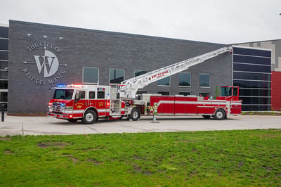 The front driver's side of an Ascendant 107' Heavy-Duty Tractor Drawn Aerial Ladder parked in front a large modern building in an urban area.