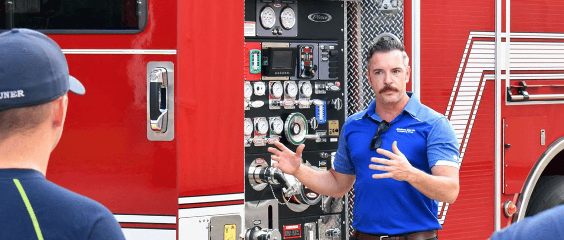 A man in a blue polo shirt talks to people while standing in front of a red fire truck.
