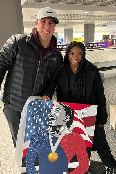 A man in a black coat with white hat holds a piece of art he created of Simone Biles at the Olympics while standing next to Simple Biles at an airport.