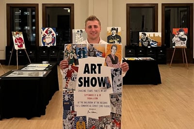 A man stands with a poster about an art show, with easels and tables filled with portrait art in the background.