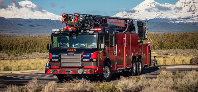 A red with black fire apparatus in a parking lot surrounded by grass and trees with mountains and a blue sky in the background.