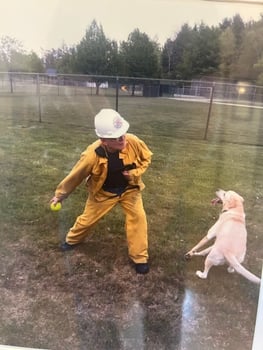 A firefighter in a yellow rain suit and white helmet throwing a tennis ball for a golden retriever in a grassy field.