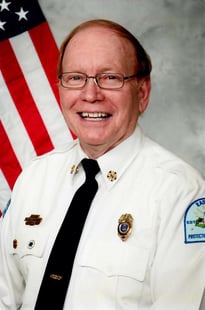 A headshot of a firefigther named Bill in dress whites with an American Flag in the background.