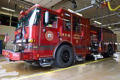 A red electric fire truck is parked in a station bay and plugged into overhead power.