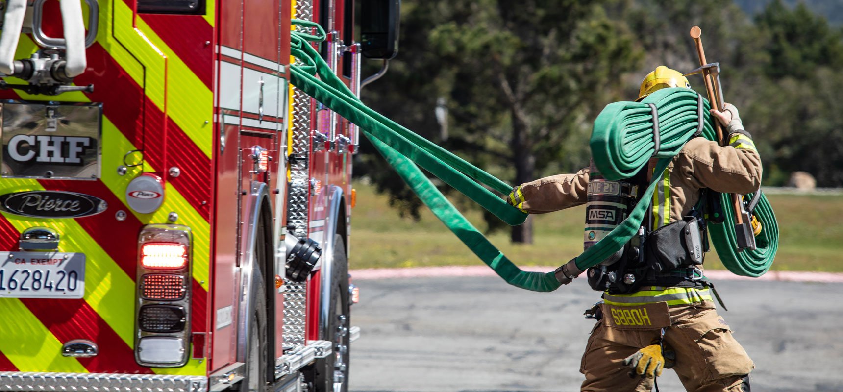 A firefighter in khaki turnout gear pulls a green firehose from a red firetruck speedlay with one hand as his other arm balances tools and a green hose pack on his shoulder. 