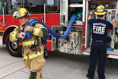 A firefighter in khaki turnout gear pulls blue fire hose from a fire truck crosslay while another firefighter operates the pump panel. 