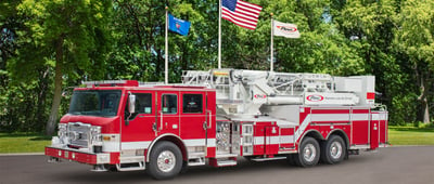 Austin Fire Department’s red Ascendant 100’ Heavy-Duty Aerial Tower fire truck is parked in front of a green tree background with three flags flying.