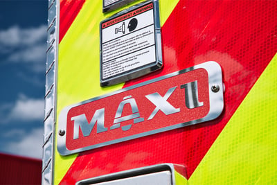 A red and stainless-steel logo reading ‘MAXI’ on a firetruck over red and yellow stripping. 