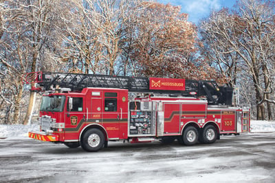 A red and black aerial fire truck with ‘Mississauga’ written on the aerial device is parked in front of a snowy background with trees. 