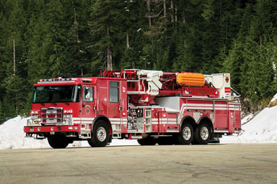 A red Pierce fire truck is parked with snow and evergreen trees in the background. 