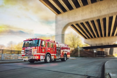 A red Pierce Volterra electric pumper fire truck drives beneath an overpass with a blurry daytime background.