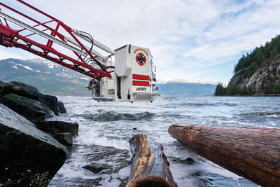 A white aerial platform is extended below grade and hovers above a rocky shore with waves and logs floating in the foreground and evergreens and mountains in the background. 