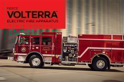 A red Pierce Volterra electric fire apparatus is driving with a blurred city background. Pierce Volterra Electric Fire apparatus is written in black on a red rectangle in the top left corner. 