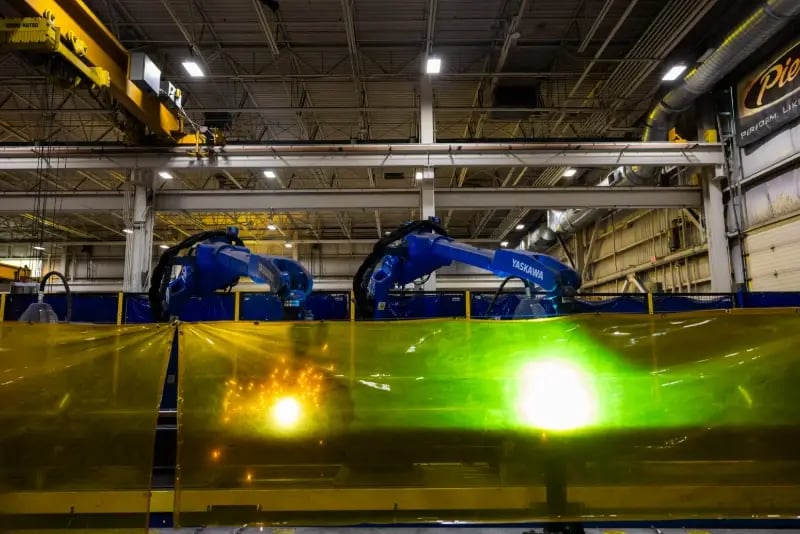 Two automated robotic welding arms in operation behind a protective screen at a Pierce Manufacturing facility.