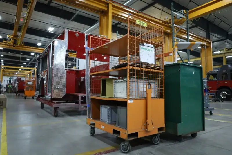 An orange parts cart in front of a red fire truck body inside a Pierce Manufacturing assembly facility.