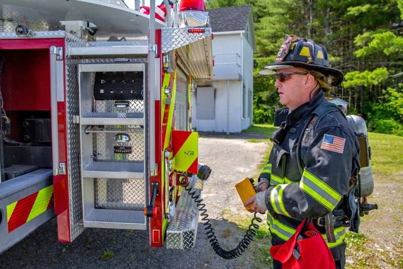 A firefighter standing on the side of a fire truck uses a control to manage the aerial stabilization system with a green wooden background.  