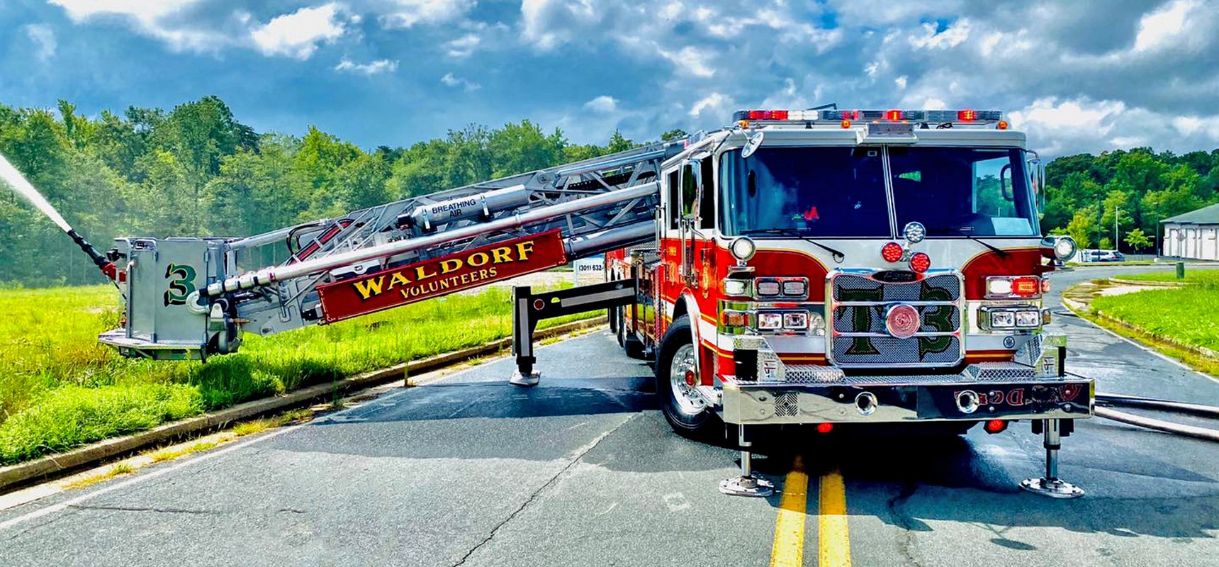 A red fire truck sits in the middle of an asphalt road with the aerial stabilizers placed and ladder extended spraying water. 