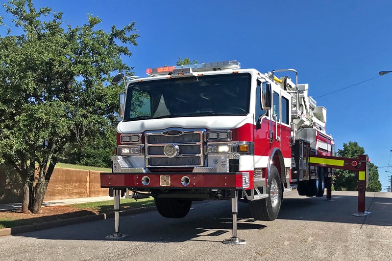 A red and white fire truck is elevated on H-Style stabilizers showing the 18-inch ground penetration and the leveling capabilities. 