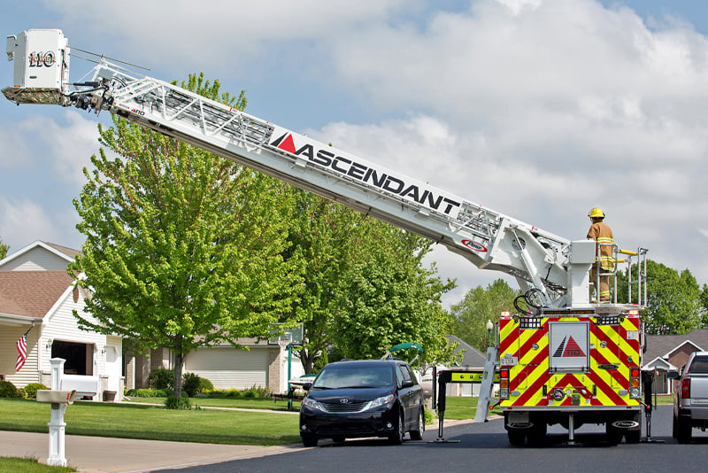A Pierce aerial is on a residential street with the ladder raised showing its ability to short jack on a narrow roadway with parked cars surrounding the apparatus. 