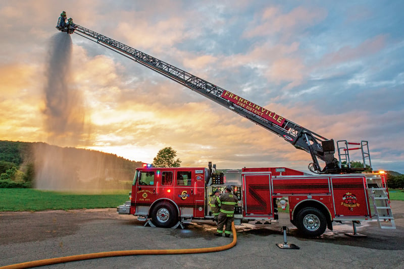 A red aerial fire truck sprays water from its tower in a country setting. 