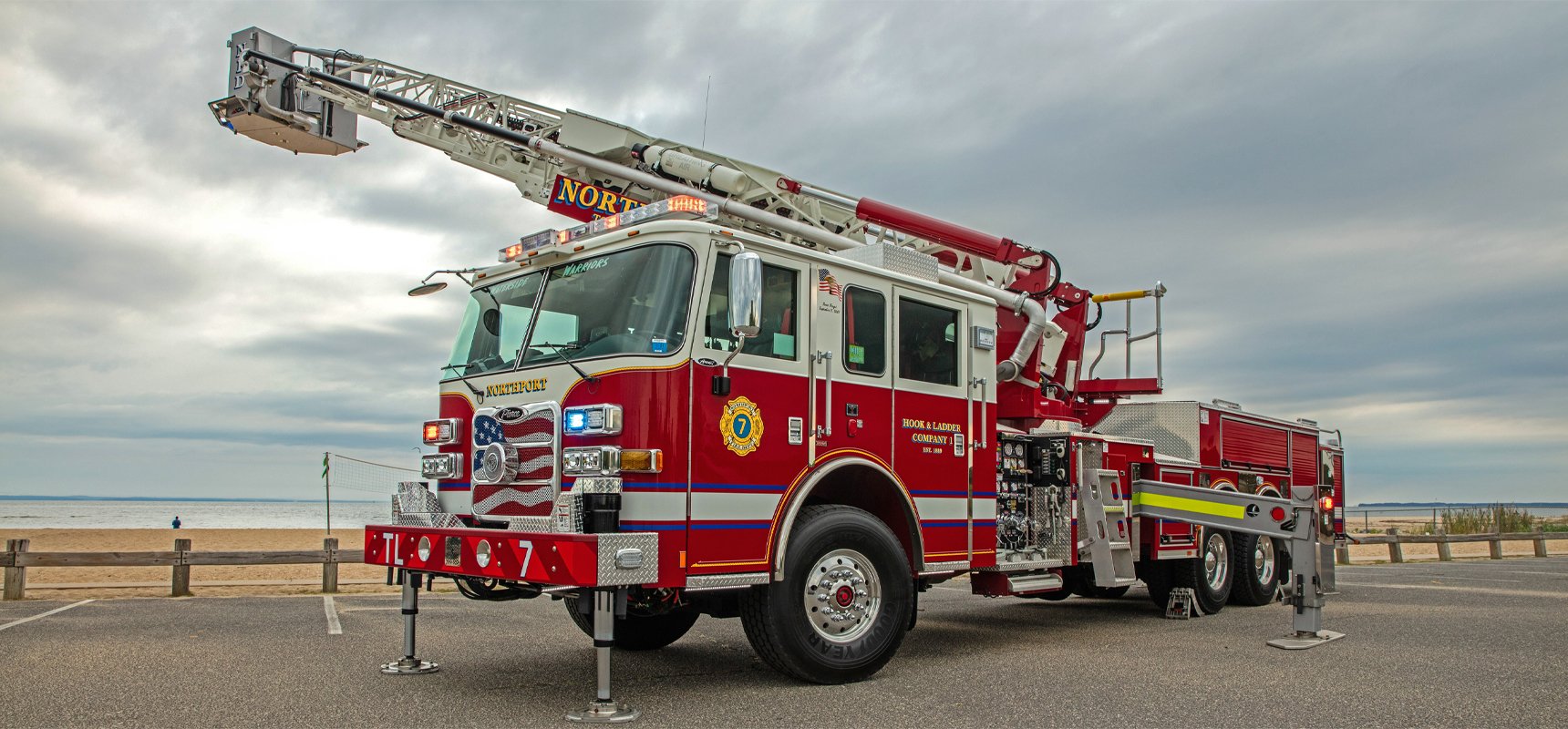 A red and white aerial fire truck with a lifted ladder is pictured on the edge of beach and body of water at dusk.