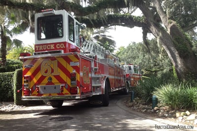 The rear end of a tiller fire truck is seen turning around a narrow driveway surrounded by green vegetation with a large southern live oak tree overhead.