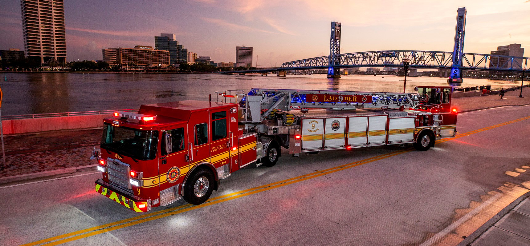 A red and white tractor drawn aerial fire truck with its emergency lights turned on is parked at sunset in front of a river and bridge landscape. 