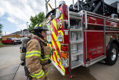A firefighter in full bunker gear walks around the rear end of a red fire truck to access the rear mount aerial top access ladder.