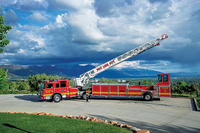 The red Salt Lake City aerial fire truck with no pump and no tank is parked with its white aerial device elevated with mountains and a blue sky filled with clouds in the background. 