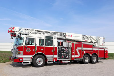 A red and white Pierce aerial fire truck with a pump and tank is parked outside a white facility with green grass and a blue sky background. 