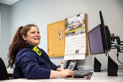 A woman with long, brown curly hair in a pony tail smiles at the camera as she works at a monitor at a desk in an office setting.