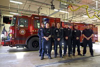 Members of The City of Madison Fire Department stand together smiling in blue, casual fire station uniforms in front of the red Pierce Volterra electric fire truck