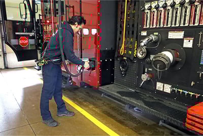 A firefighter in a blue, casual station uniform plugs an electric fire truck into an overhead charging cable inside a fire station. 