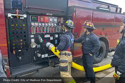 Three firefighters stand at the pump operator panel on a red Pierce Volterra electric fire truck managing controls on scene.