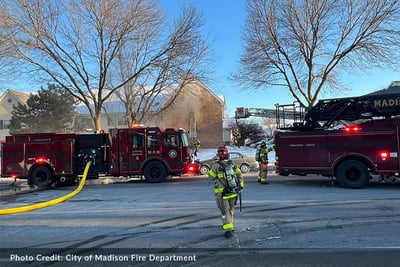 A firefighter in full gear walks on the street toward two red Pierce apparatus operating on scene.