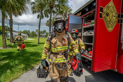 : A fire fighter in full gear walking next to a red fire truck with palm trees in the background.